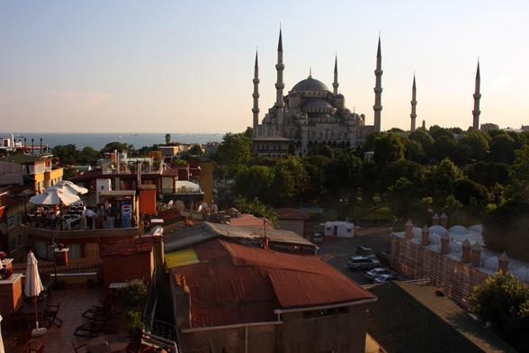 The sun is setting so the image of the Blue Mosque in Istanbul, Turkey is mostly a silhouette of its minarets and dome. The sky is a pale yellow/orange with barely a tinge of blue. The red roofs of businesses  are below in the foreground.