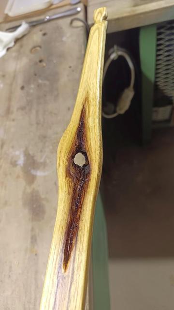 Wir blicken auf den unteren Wurfarm, der mittig ein Loch von einem Ast hat und durch die Witterung und Wetter, das Holz im Inneren des Stammes auch verrottet ist und dadurch ein längliches Loch gebildet hat. Das Holz ringsum ist Honiggelb und das Loch am Rand dunkelbraun