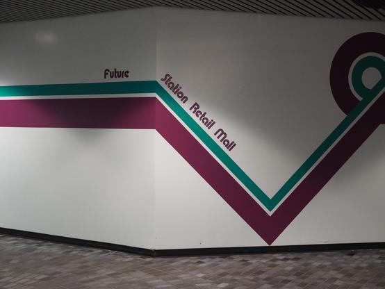 A white wall painted with a maroon and teal stripe on it. As the stripe goes around a corner, it dips 45 degrees, then rises again, doing a loop near the ceiling. If you look close, there is a shadow under the wall cast across the varied tones of blue-grey tile. This corridor has the striped polished metal ceiling typical of Bay and Corona LRT stations.

Oddly spaced, and spanning the corner in a thick weirdly rounded font that is very Edmonton in the 1980s, printed in matching maroon is the words "Future Station Retail Mall.