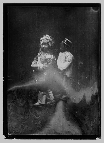 The image depicts a black and white photograph of two children standing on what appears to be an uneven, possibly rocky surface. The child in the foreground is wearing traditional Native American-style clothing including moccasins with fringes, leggings, and a decorated headdress that resembles a snowman or winter scene made up of small objects like beads or seeds. This attire suggests cultural significance, perhaps for dance or ceremony.

The second child stands behind slightly to their right side in the background. They are dressed more conventionally in Western-style clothing with short sleeves and what seems to be light-colored trousers; they wear a hat similar to those worn by Boy Scouts during that era. Their stance is relaxed compared to the child beside them, who appears poised.

There's an aura of stillness or pause as if caught mid-step on their walk down Chinatown streets in San Francisco between 1896 and 1906 based on contextual information given about this image. The black-and-white nature captures a historical moment without color distractions, focusing the viewer’s attention on texture contrasts such as clothing details versus rough ground surface.

The photo's age is indicated by its faded quality and graininess which suggests it has been preserved for over a century from that time period. This snapshot of history offers insight into cultural diversity wit [...]