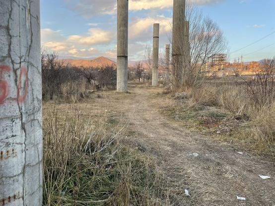 Pillars of an unfinished viaduct in Spitak. Grass in the foreground, mountains and a factory in the background.