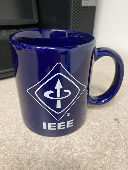 Eine dunkelblaue Tasse. Darauf ist in weiß das IEEE Logo. Das Logo ist viereckig in Anspielung auf Benjamin Franklins Drachen. Im Innern des Drachen ist ein Pfeil der nach oben zeigt und ein weiterer Pfeil,  der sich gegen den Uhrzeigersinn um den ersten Pfeil windet. Dies symbolisiert die Rechte-Hand Regel für die Beziehung zwischen Strom und Magnetfeld.