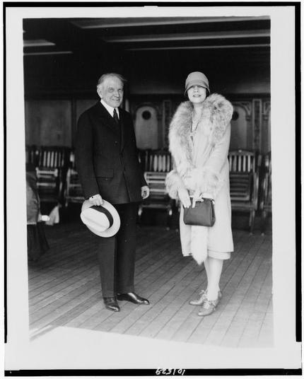 The image is a black-and-white photograph of two individuals, a man and a woman, standing on what appears to be the deck of a ship. The man is dressed in a formal suit with a white shirt and a dark tie, holding a white hat in his right hand. He is wearing dark shoes and has a confident posture. The woman is dressed in a light-colored coat with a fur-trimmed collar, a light-colored hat, and a dark purse. She is also wearing a dress underneath the coat and has a necklace and earrings. The deck beneath them has wooden planks, and there are wooden chairs and a railing visible in the background. The image has a white border with a black line around it, and there is a number "623101" at the bottom right corner. The setting suggests a formal or elegant occasion, possibly during the early to mid-20th century.