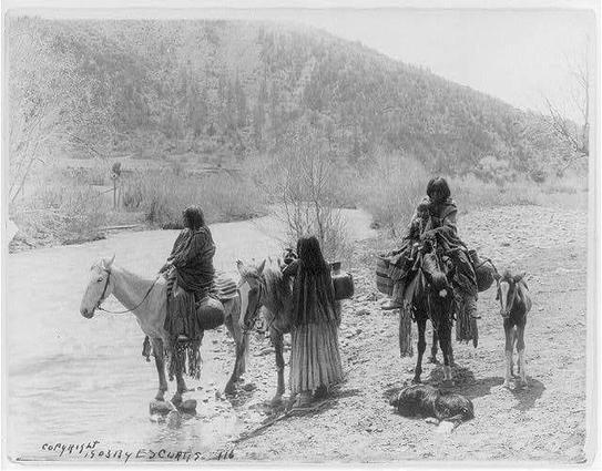 The image depicts three Apaches with horses, one of which has a child on its back. They are stopped beside a stream in an outdoor setting, possibly during the early 1900s based on historical context and clothing style. The photograph is monochromatic, suggesting it's black-and-white or sepia-toned. A dog can be seen near the horses' feet, and water jugs indicate they may have been traveling to fetch water for their journey.