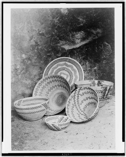 This black and white photograph shows a collection of woven baskets with various patterns on their rims. The background appears to be an outdoor setting, possibly the exterior of a building or cave-like structure, given its rough texture and dark tones. There are seven bowls in total; some stand upright while others lie partially collapsed at different angles. Each basket displays intricate geometric designs such as zigzags, diamonds, triangles, concentric circles with smaller rings within them, and other symmetrical patterns that suggest cultural significance or craftsmanship associated with their creation.