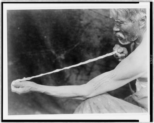 A black and white photograph showing an older man with a rough, weathered appearance holding what appears to be a string or rope. His face is smeared with dirt or soot, adding to his unkempt look. The most striking feature of the image is the numerous tattoos etched onto both arms in neat rows containing various characters that may represent writing from an indigenous language or symbols specific to Native American culture.

The man's right arm displays a series of dots and lines forming words which are difficult to decipher due to their size, while his left arm features longer strings of text. The tattoos extend across the full length of both arms, with some markings more prominent than others. His eyes are closed or nearly shut as if in deep concentration or fatigue.

The background is blurry but suggests a natural outdoor setting, possibly forested terrain given its dark and indistinct appearance. This image seems to document aspects of Native American culture during the early 20th century, with an emphasis on body art through tattoos, which could have had cultural significance at that time.