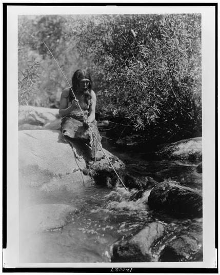 The image is a black and white photograph depicting an indigenous person, likely from the Miwok tribe, as indicated by the reference in the provided information. The individual appears to be sitting on a large rock beside a flowing creek or river. They are holding what seems to be a spear or fishing rod with one hand while grasping something that could possibly be food with their other hand.
The person is wearing minimal clothing and has long hair, which suggests they might belong to an earlier period in history when such attire was common among indigenous tribes in the region. The surroundings include dense vegetation and various rocks along the water's edge, creating a natural setting typical of Native American landscapes during that time. This photograph captures a moment of quietude or daily activity within this person’s life as they engage with their environment.
The image is credited to Edward S. Curtis (1868-1952), an American photographer known for his documentation and portrayal of various indigenous peoples in the United States through photographs, which were published under titles such "American Nudes" and books like "The North American Indian," among others that sought to preserve cultural heritage as well as artistic expression. The photograph is noted with a reference code from Loeper Collection (LZ-20371) on its bottom right corner.