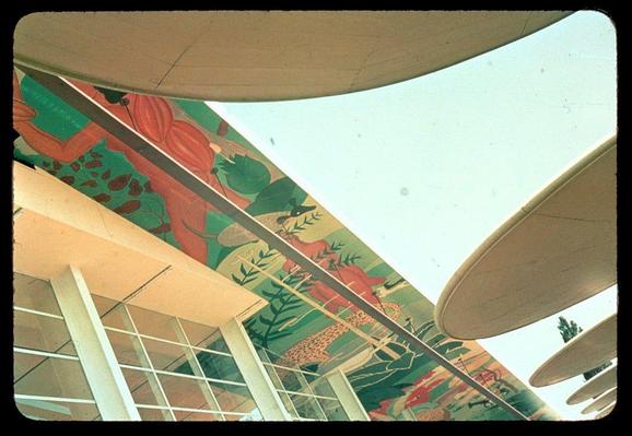 The image is a photograph of a colorful mural painted on the exterior wall and ceiling structure of an architectural building. The art features vibrant, tropical plants with shades of reds, greens, oranges, and yellows set against a green background. A clear sky can be seen in the upper part of the photo through large windows or openings within the architecture.
The photograph is taken from below looking upwards towards the mural and ceiling structure, giving an angled perspective on this artwork. The building appears to have modernist architectural elements with glass panels and white beams supporting the overhanging roof-like structures. There are no people visible in the image; it focuses solely on the art piece and part of its surroundings.
The additional information provided suggests that the mural was created for a World's Fair, possibly related to an exhibition involving Ecuador during either 1939 or 1940. Gottscho-Schleisner, Inc., is mentioned as likely being responsible for producing this photograph slide. The source of the image indicates it belongs to Loener collection and can be found on their website at <https://images.loener.nl/gottscho-schleisner/full/691e/691ef6d5450e57a2674f7aa9.jpg>.