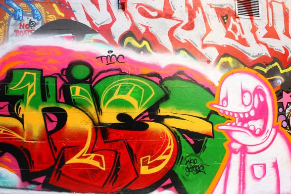 Very bright and vibrant graffiti artwork in Hosier Lane. It is painted in an alley that has graffiti artwork covering both sides of the alley and more. This one has lettering and a cartoon character with a big mouth and teeth plus big, oval eyes. Colors include hot pink, green, red, orange, yellow and a little blue. Hosier Lane, Melbourne, Australia