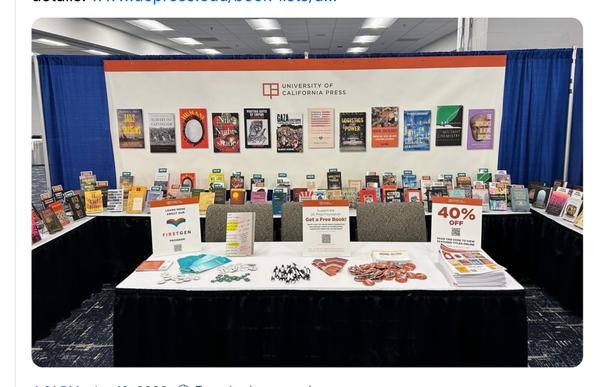 A gorgeous University of California Press book display: the background wall is a white banner with a row of book covers, in a pleasingly jagged horizontal line. On the three sides of the table around the booth are rows of books, some with labels sticking out. At the front is a table, three chairs behind it, and various announcement cards on the desk, together with lots of GORGEOUS SWAG (pens, bookmarks, badges) and printed conference catalogues.