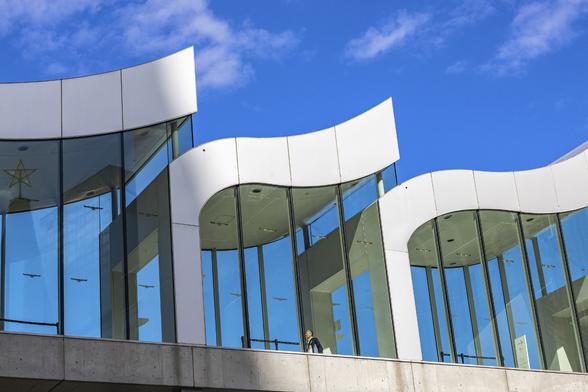 A man is going through a glass pathway covered with wave-like white shapes under a bright blue sky in Osaka, Japan. A few light little clouds are ascending the sky in a movement reminding of the building shapes. The man is lit in the sun while half of the pathway is in the shadows.