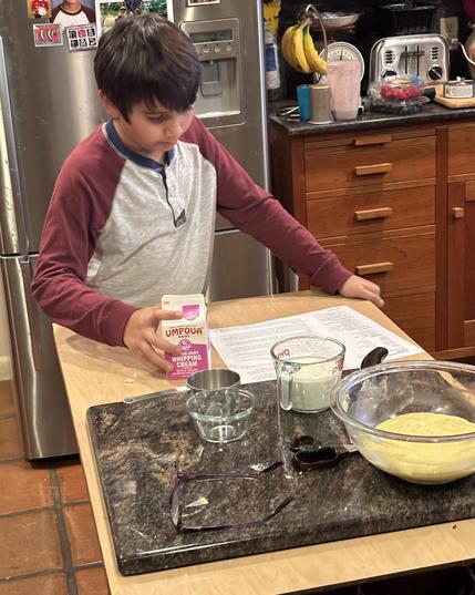 A child prepares ingredients in a kitchen, holding a carton of whipping cream. A recipe is laid out on the counter, along with bowls of various ingredients and measuring tools. In the background, there are kitchen appliances and bananas on display.