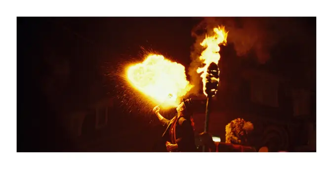 A colour photograph of a circus performer in clown Makeup standing on a ladder breathing fire. A huge fireball emanates from their face and illuminates the crowd, watching in the foreground, a man on stilts carrying a torch.