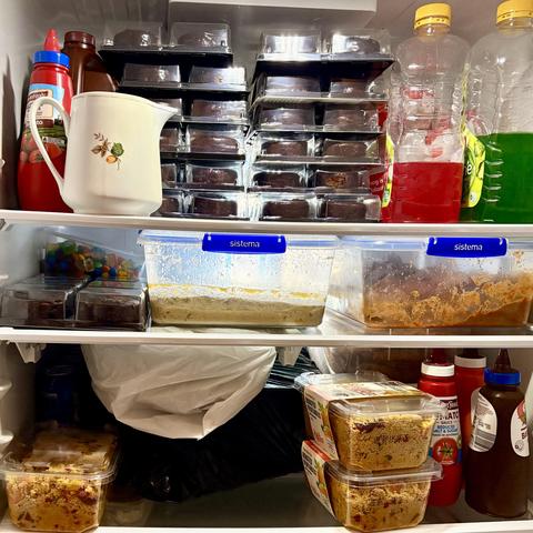 A refrigerator holds various food items, including containers of masaman and vegie curries, stacks of mini chocolate mouse, and bottles of ketchup and sauce. The shelves are organized with labeled containers and plastic bags.