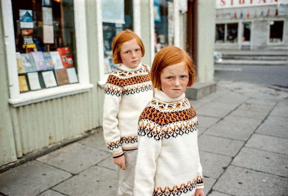 The image shows two young children with reddish hair, standing on a sidewalk. They are wearing traditional Scandinavian sweaters known as "Lusekofte," which feature intricate patterns in white and shades of orange-brown, possibly indicating that the photo is from Norway or another Nordic country. The child closest to the camera appears slightly more intense than their companion, with furrowed brows and a direct gaze into the lens.
The background reveals an urban setting; there's a storefront on the left side displaying various items for sale through its window. Notable features include signs that suggest commercial activity in the area. There is also a staircase leading up to another building or establishment visible in the distance, along with more shops and possibly residential buildings further away.
The street appears quiet without much pedestrian traffic at this moment captured by the photographer Ed van de Elsken as per the reference provided: "Verliefd op de fotografie van Ed van de Elsken - NRC." The overall mood of the image seems candid, perhaps taken during a casual day out or an ordinary walk in their neighborhood.