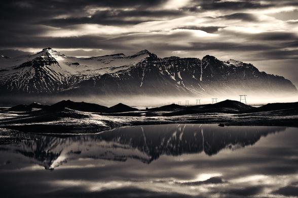 a large coastal mountain range, topped with snow and ice and bathed in speckled morning light and opposing shadow, while sea mists drift in from the rough seas... in the foreground, small foothills and deep pools of water create a near perfect mirror