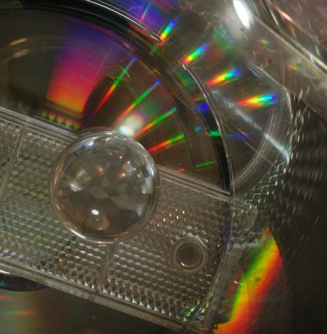 The same reflector was placed on a CD, and the small glass sphere was placed on top of the CD. A glass cylinder without a base was placed on the CD and reflector. The whole assembly was illuminated with white LED light, and due to the refraction of light caused by the CD, many spectral colours appeared. The cylinder reflects the reflector in a distorted way.