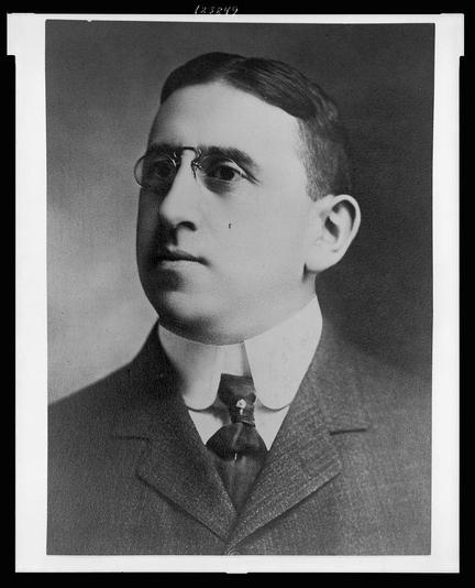 The image is a black-and-white portrait of a man from what appears to be the early to mid-20th century. The man is wearing a formal suit with a white shirt and a dark tie. His hair is neatly combed back, and he is wearing round, wire-rimmed glasses. The background is plain and dark, which helps to focus attention on the subject. The photograph has a rectangular frame with a white border, and there is a small number "(23849)" in the top right corner, possibly indicating a catalog or reference number. The man's expression is neutral, and he is looking slightly to the left of the camera.