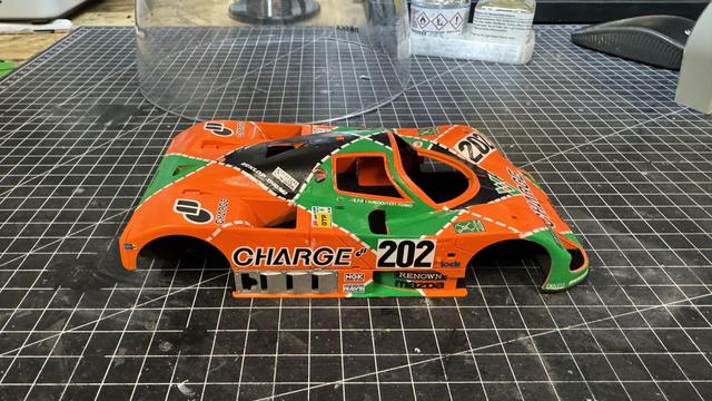 Side profile view of completed Mazda Charge 767B 1:24 model showing the full orange and green racing livery, number 202, sponsor decals, and assembled body panels on a black grid mat