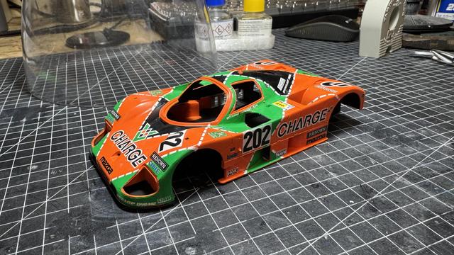 Side profile view of completed Mazda Charge 767B 1:24 model showing the full orange and green racing livery, number 202, sponsor decals, and assembled body panels on a black grid mat