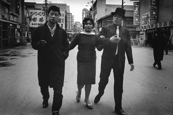 The image is a black and white photograph depicting three individuals walking down an urban street. On the left, there's a man in dark clothing with a serious expression, holding what appears to be a cigarette between his fingers. In the center stands another woman wearing a long-sleeved dress with a high collar; she has her arms crossed over her chest and seems focused on something out of frame. The third individual is a formally dressed man sporting a black hat, white shirt, dark tie, suspenders, and holding what looks like a cigarette in his left hand while carrying an object under his right arm that's not clearly visible due to the angle. They all seem engaged with their surroundings rather than each other.

The street behind them features various storefronts with Japanese characters on signs, indicating this scene likely takes place in Japan or is related to Japanese culture. The architecture and style of signage suggest it could be a busy commercial area from an earlier era. There are faint silhouettes of passersby walking along the sidewalk, adding depth to the street's bustling atmosphere.

The image has a candid quality, capturing a slice of life moment with a grainy texture typical for film photography taken in daylight conditions during mid-20th-century urban environments.