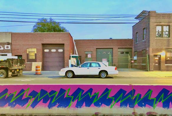 Colorful green, pink and purple graffiti lines a concrete barrier separating a bike lane from the main road in Astoria, Queens. A white sedan sits idly along the street. The buildings are low, one and story brick buildings.