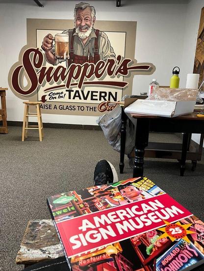 A copy of the American Sign Museum book, with its cover dominated by colourful illuminated signs, is visible in the foreground, resting on someone's knee and their foot poking out from behind the book. In the distance is a white wall with a mural painted on it. The mural depicts a kindly grey-haired man holding a glass tankard of beer with lettering underneath that reads "Snapper's Tavern, Come on In!, Raise a Glass to the Craft".