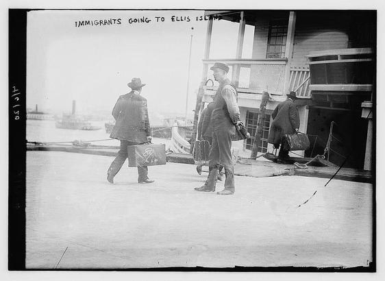 The image is a black-and-white photograph depicting a scene from the early 20th century. It shows a group of individuals, likely immigrants, walking towards a building with a porch. The setting appears to be near a body of water, as there are boats visible in the background, suggesting a harbor or dock area. 

The individuals are dressed in attire typical of the era, with hats, suits, and boots. One person is carrying a suitcase, which has a handle and appears to be a standard suitcase of the time. The building they are approaching has a wooden structure with a railing and what looks like a door or entrance. The photograph is labeled at the top with the text "IMMIGRANTS GOING TO ELLIS ISLAND," indicating that this scene is related to the immigration process at Ellis Island, a historic U.S. immigration center. The image conveys a sense of movement and transition, as the individuals are embarking on a significant journey.