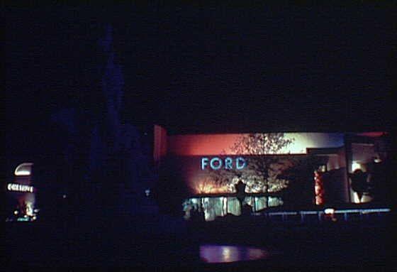 The image shows a night scene at an exhibition, likely from the 1939 or 1940 World's Fair. A prominent sign reads "Ford," indicating it is associated with Ford Motor Company. The building has red and blue lighting that outlines its shape and features decorative elements like trees illuminated in white lights against a dark background. Other structures are visible, including one labeled "Columbia" on the left side of the image. There appears to be some activity or movement within the venue as silhouettes can be seen through the windows. The overall ambiance is that of an exhibition with architectural displays and lighting meant for nighttime visitors.
