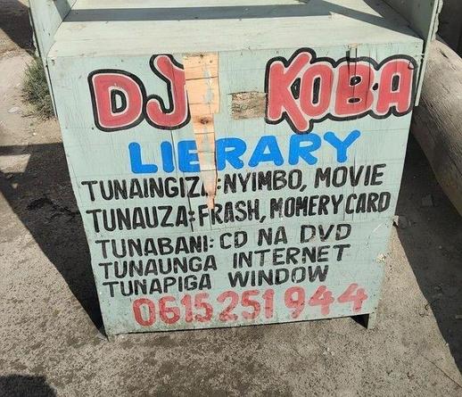 Street stall with painted lettering on the side that reads: "DJ Koba, Library, Tunaingiza: Nyimbo, Movie, Tunauza: Frash Momery Card, Tunabani: CD NA DVD, Tunaunga Internet, Tunapiga Window, 0615 251 944".