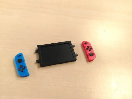 Ein 3D gedrucktes Nintendo Switch 1 Modell, ca. 4,5 x 2cm groß mit einem roten und einem blauen Controller. Die Controller sind abgesteckt und liegen neben der Basis. Man sieht, dass aus den Seiten je zwei kleine Stifte ragen auf die die Controller offenbar mit passenden Löchern gesteckt werden.