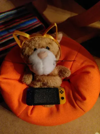 Eine süße Nicci-Katze auf einem orangenen Handysitzsack. Sie ist braun gestreift, trägt ein paar 3D gedruckte Katzenohren und hält auf dem Schoß eine 3D gedruckte Nintendo Switch in passender Größe. Die Switch hat ganz stilecht einen orangenen einen violetten Controller, die Tasten sind schwarz und auf dem orangenen Controller gut erkennbar. Der Mittelteil ist aus schwarzem Glitzerfilament für einen Bildschirm-artigen Effekt.