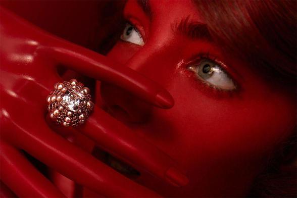 A close-up of a shiny red hand with a decorative ring adorned with pearls and intricate designs, partially covering a red face.