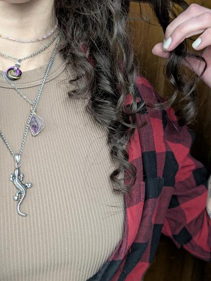 Kat taking a close up selfie. They're wearing a tan ribbed crop top and a red and black flannel. Their necklace stack today is the classic salamander and amethyst combination paired with their always on nonbinary pendant and sapphic choker. They're holding up some of their curls to show how their hair has held up over the past couple of days. A little messy, but retaining their coils.