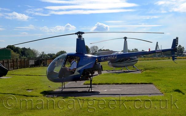 Side view of a small, metallic blue helicopter parked facing to the left on a concrete landing pad surrounded by grass.
The copter has it's doors missing, and a man in a white shirt can be seen sitting in the left seat, with the person on the right in shadow.
There is a silver lightning flash on the side of the fuselage, with silver text "R22 Beta" on the upper rear fuselage, and the silver registration "G-HIZZ" on the long, thin, tail boom.
Green grass fills the foreground and a large part of the background, with another, similar helicopter behind this one, and trees and a grey hangar behind a wooden fence.
Blue sky with occasional streak of cloud fill the rest of the frame.