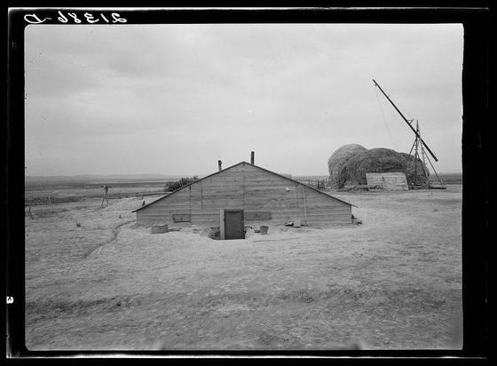 The image depicts a rural farm setting in black and white. A large cylindrical silo is positioned on the left side of the frame, with its entrance facing away from us. To the right stands an old wooden barn-like structure with two visible doors: one appears to be closed while the other seems open. The building exhibits weathered siding and a pitched roof, indicative of considerable age or exposure to harsh conditions.
In front of this building lies a vast expanse of dry grassland that extends into the distance under an overcast sky. On the right side in the background stands another agricultural implement with tall stilts supporting what appears to be hay bales secured on top, suggesting recent harvest activity.
The ground is largely barren except for scattered patches of vegetation and some farm equipment or objects lying idle near the barn structure's foundation. The overall scene conveys a sense of solitude and hard work associated with rural life during an earlier era based on architectural styles and absence of modern machinery.