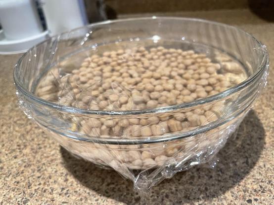 Chickpeas soaking in water in a medium size glass bowl, covered with cling wrap.