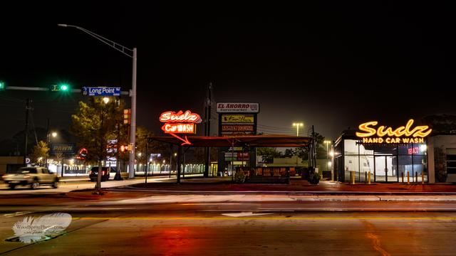 Early morning street scene on Long Point Road in Houston before sunrise, with thick fog softening neon car wash signs and green traffic lights. Dry pavement, empty lanes, and storefronts fading into mist.