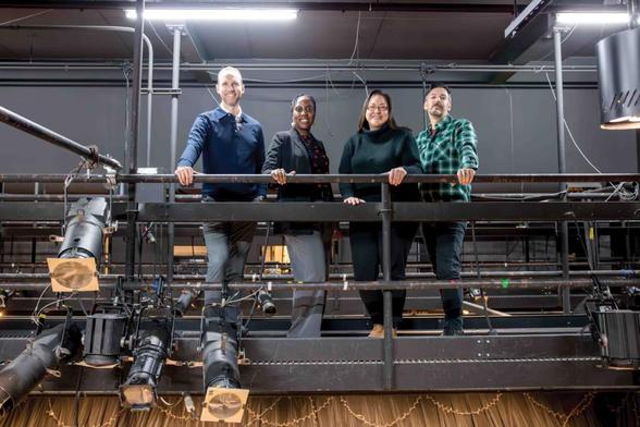Photo taken inside a theatre of four people smiling and posing together on a balcony above the lighting rig.