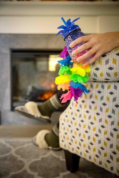 Photo taken in someone's living room of the side of an armchair, inhabited by someone wearing slippers and a flower necklace, drinking out of a pineapple shaped cocktail jar.