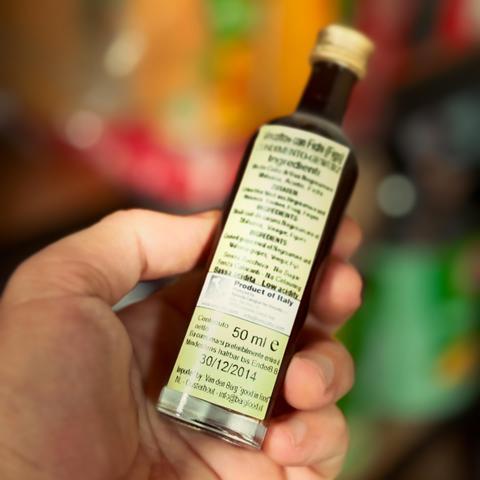 A person holding a small brown bottle of vincotto con fichi with a pale green label. The label has black text including a use by date over a decade ago