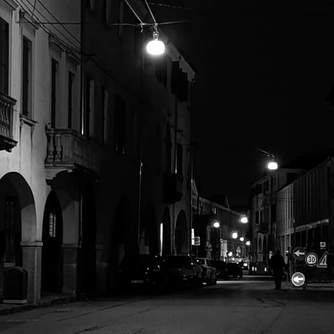 Lampioni sospesi illuminano una strada buia, tracciando un percorso. Sul lato sinistro portici di vecchi palazzi. Foto in bianco e nero.
