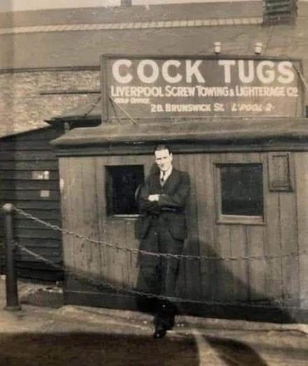 A suited man stands confidently in front of a weathered wooden building bearing a bold maritime sign: “COCK TUGS – Liverpool Screw Towing & Lighterage Co.” The vintage black-and-white photo captures mid-20th-century industrial pride, with the man’s crossed arms and slight lean suggesting ownership or authority. The sign’s phrasing, though earnest in its era, adds a layer of unintended humor for modern viewers.