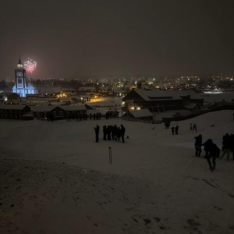 Det er et mørkt bilde fra nyttårsaften 2025. Til venstre en opplyst Røros kirke med et rødt og hvit fyrverkeri bak. Det skimtes folk som står i en snødekt Slagghaugan på Røros.