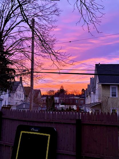 A morning sky glows with orange, pink, and soft purple above the residential neighborhood, seen from the backyard over a wooden fence, where a black rebounder leans against the fence.