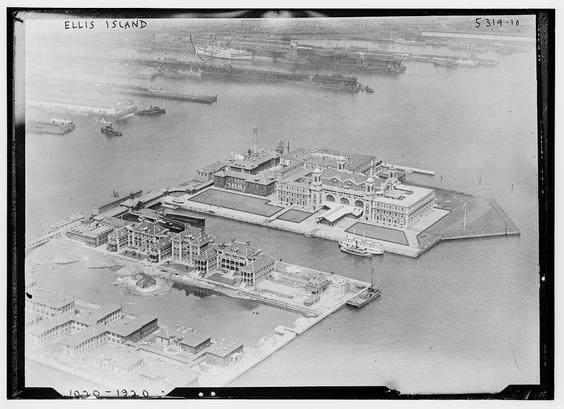 The image is a historical black-and-white aerial photograph of Ellis Island, located in New York Harbor. The photograph shows the island in its entirety, with the main immigration processing facilities prominently displayed in the center. The architecture includes a large, rectangular building with multiple wings and a central dome-like structure, which is the main immigration station. The surrounding area features a dock with a pier extending into the water, where a small vessel is docked. The island is surrounded by water, and there are additional smaller buildings and structures visible on the island, including what appears to be a hospital or other administrative buildings. In the background, there are other ships and docks, indicating a busy port area. The photograph is labeled "ELLIS ISLAND" at the top, and there are some numbers and markings at the bottom, possibly indicating a catalog or reference number. The overall scene reflects the bustling activity associated with immigration during the early 20th century.