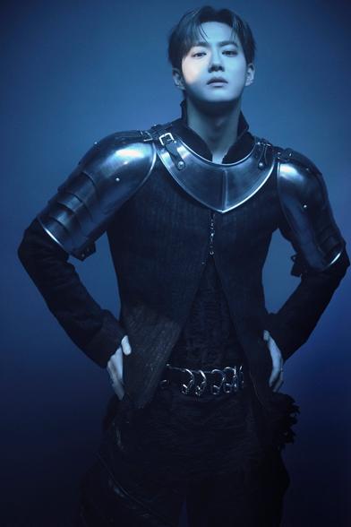Suho tilts his chin up as he stares down into the camera, wearing dark, form-fitting woven clothing and metal shoulder, bicep, and neck armor like a medieval knight