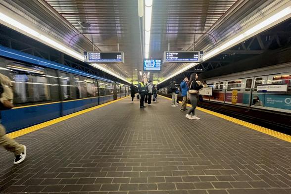 La imagen muestra una estación de metro con dos trenes y pasajeros esperando en el andén. Se observa una señal con indicaciones de destino y tiempo de llegada.