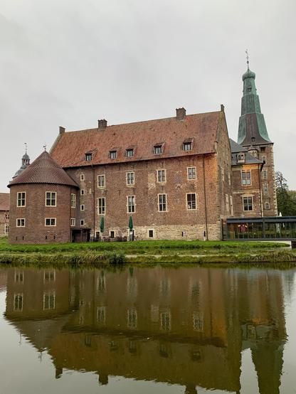 A large, traditional brick building with a steeply pitched roof and a prominent tower with a green spire reflected in a still body of water in the foreground. The building features multiple small, rectangular windows and a slightly arched entrance. There is a small modern glass extension on the right side of the building, and several closed green umbrellas are visible beside the structure. The sky is overcast.