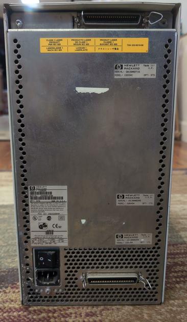 The back of a tower computer case. At the very top and very bottom are Centronix connectors. A rocker switch and IEC power inlet are at the bottom left. Most of the back of the case is bare metal. There is perforation at the bottom and around the sides for air flow. A yellow laser warning sticker is at the top and there are smaller HP logo stickers on the case, too.